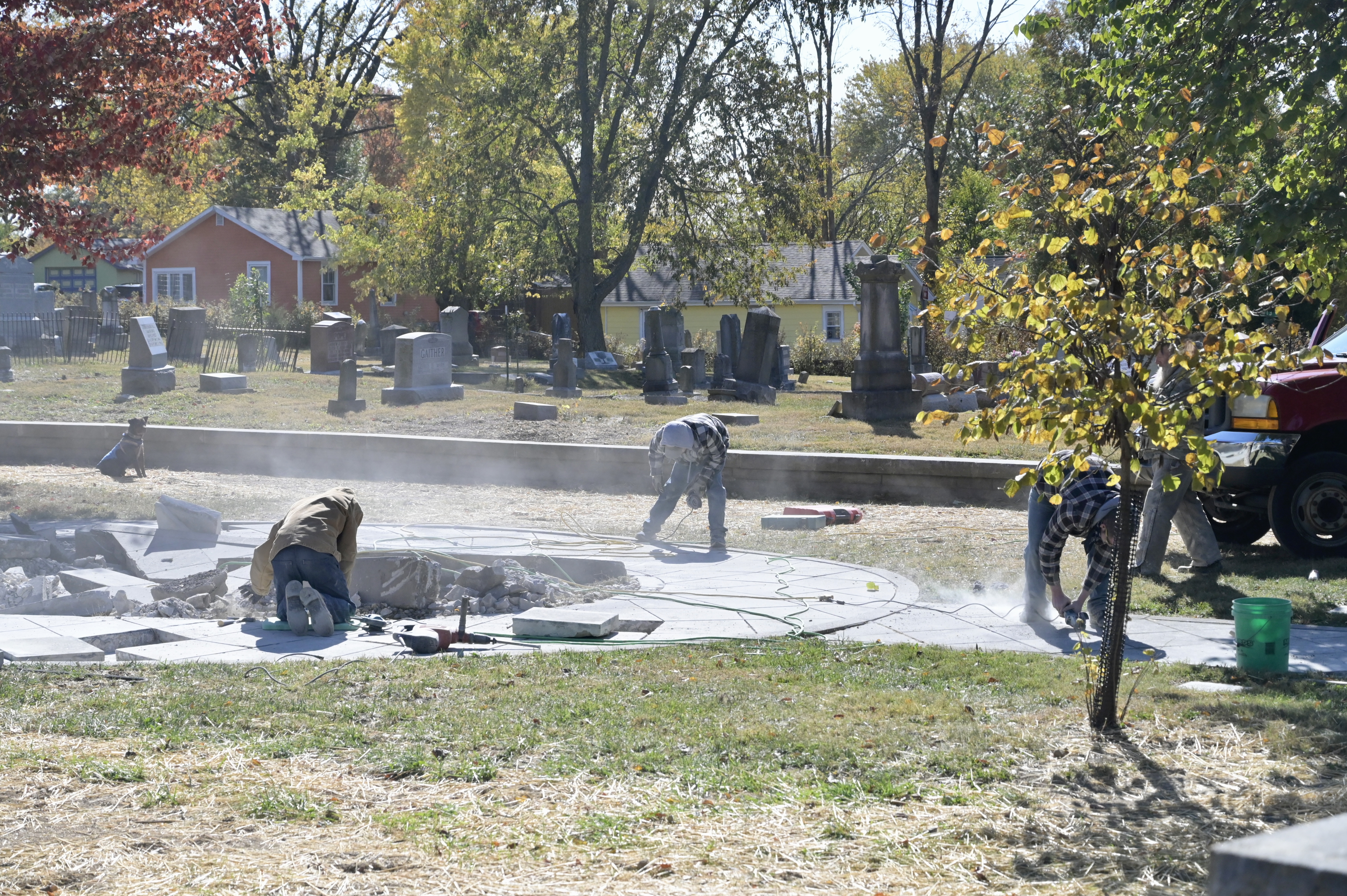 Photo shows three workers in the Rose Hill Cemetery surrounded by gravestones, working with tools to break up an old concrete foundation.