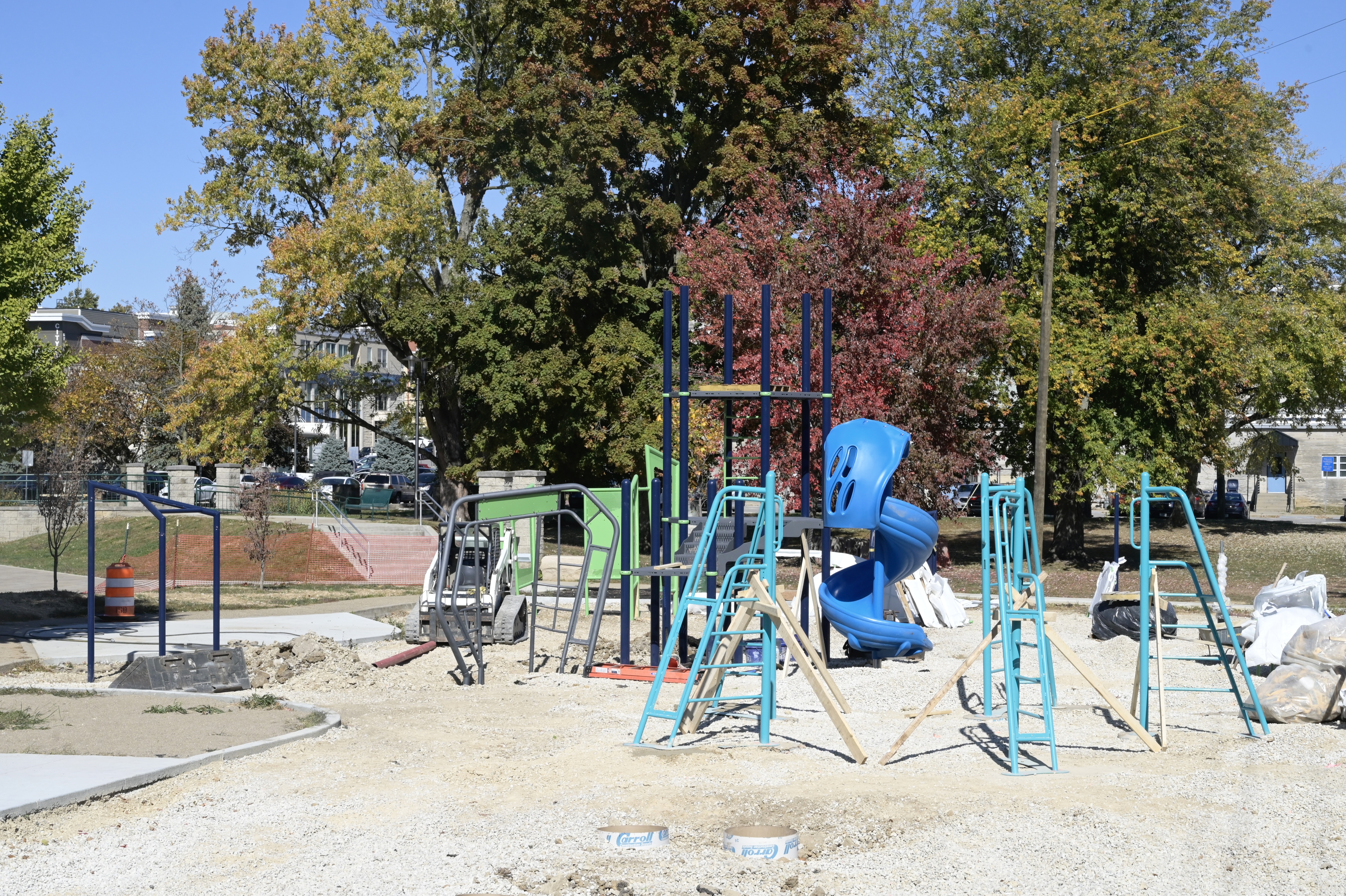 Photo shows turquoise, blue, and green structures standing in front of colorful fall trees in the bare lot where the new playground at the Waldron, Hill and Buskirk Park will be installed. 
