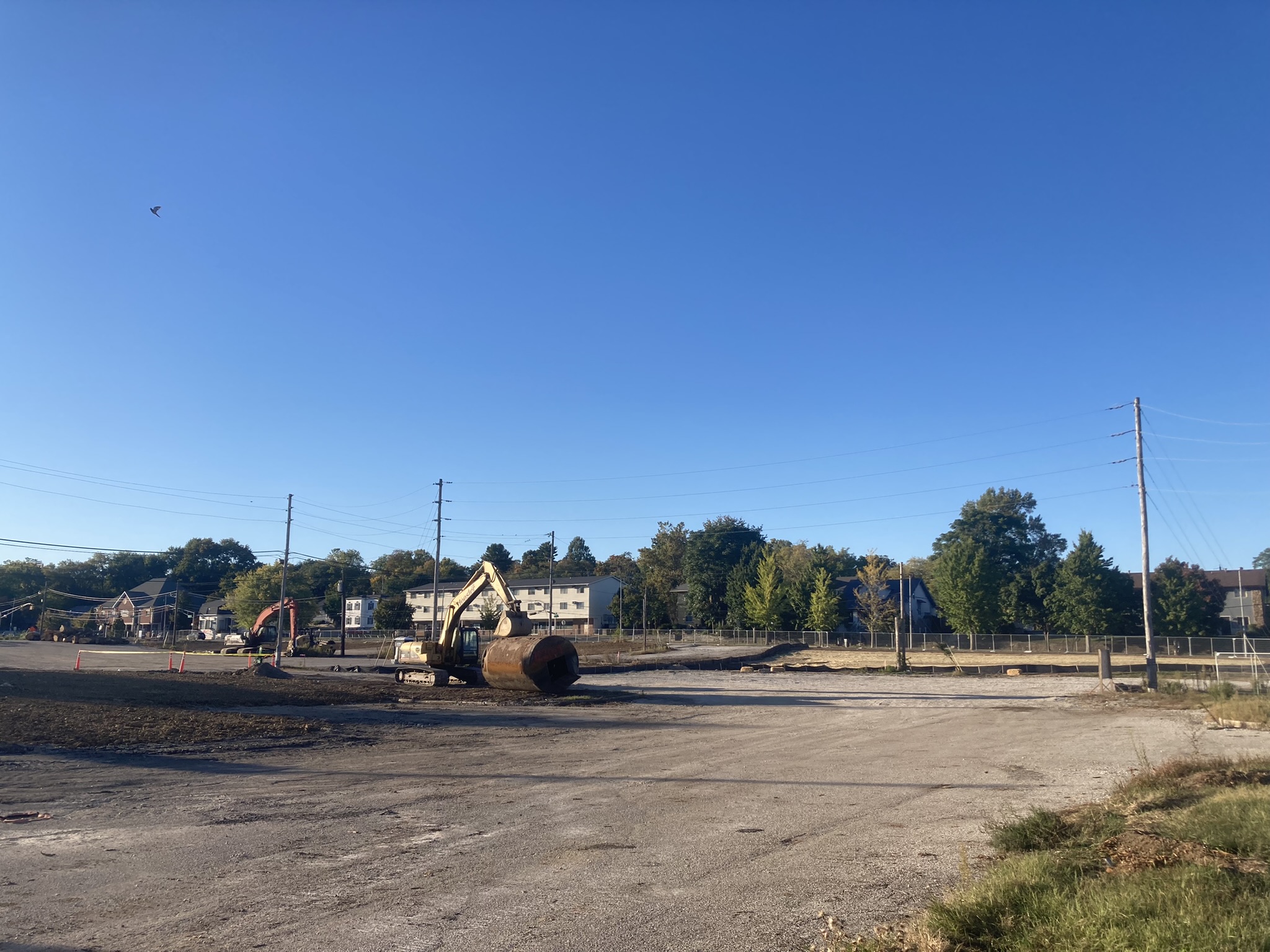 An excavator is in a cleared and fenced construction area;  its bucket rests on a cylindrical tank.  In the background, telephone poles and wires, trees and residential buildings (or small offices). 