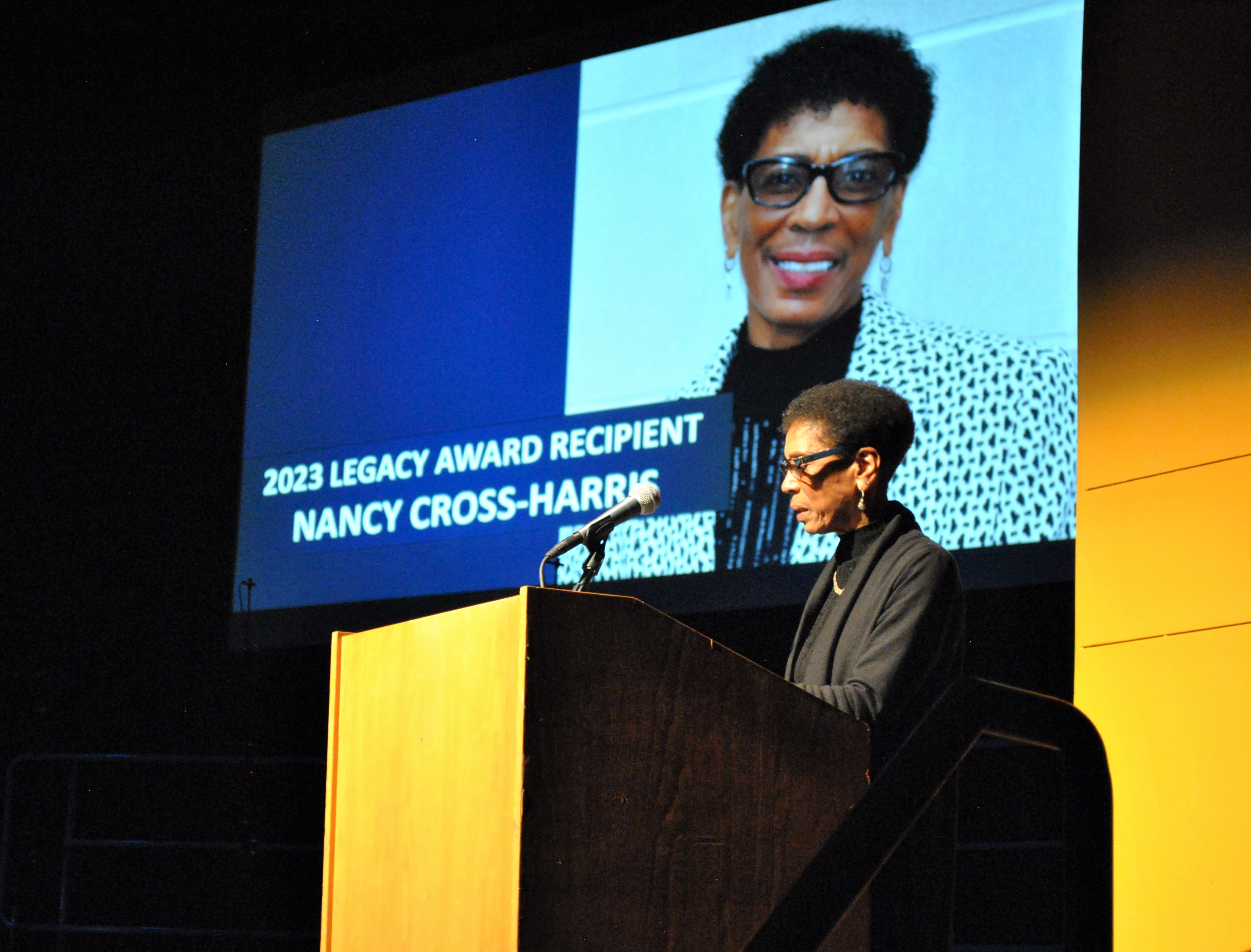 Nancy Cross-Harris stands at the podium at the Buskirk-Chumley Theater in front of a photo of herself displayed on a slideshow screen. She is giving her acceptance speech for the 2023 Legacy Award.