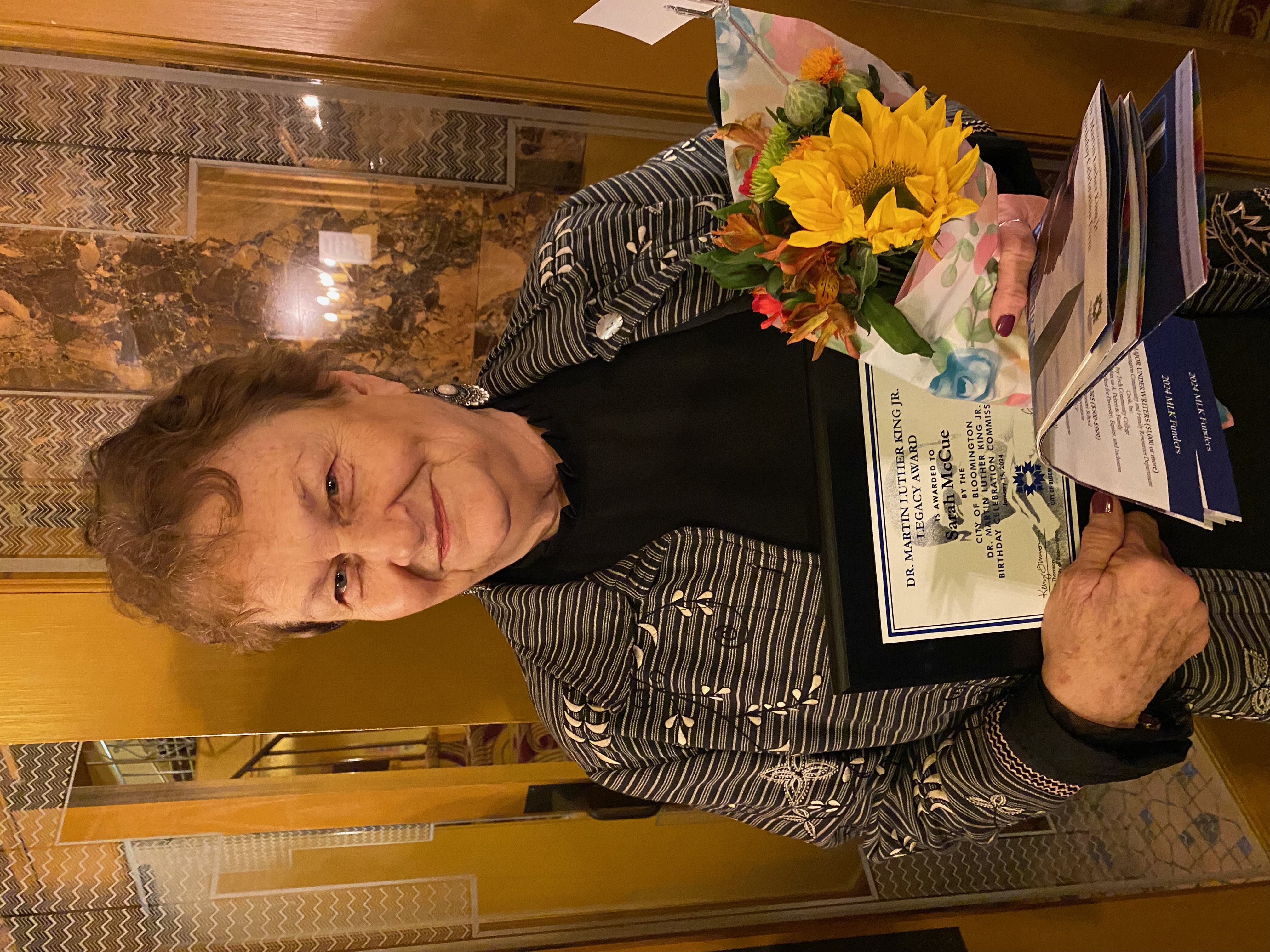 Sarah McCue holds flowers and her legacy award plaque in front of the guilded gold doors of the Buskirk Chumley Theater's inner lobby.