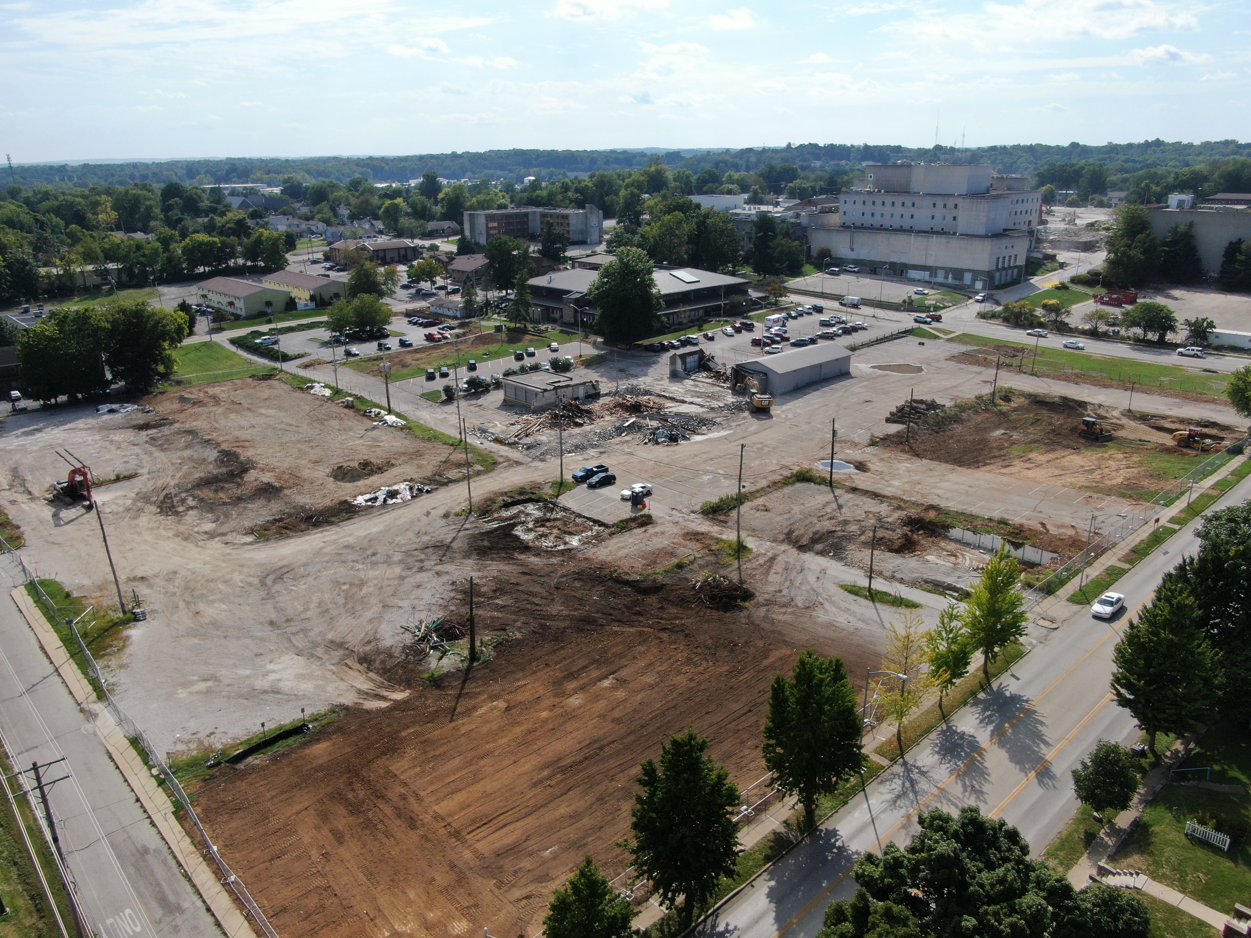 The right photo is looking southwest from the intersection of 2nd and Morton Street, there is a cleared site with exposed earth and the same equipment as the left photo working in the middle ground, the legacy hospital is shown in the background. 