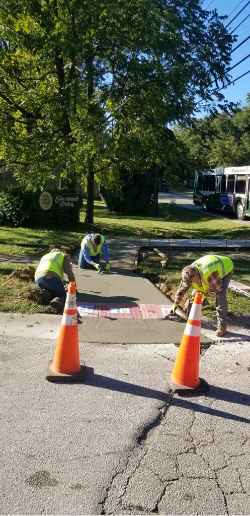 Three city workers wearing reflective safety vests install an ADA compliant sidewalk ramp on S Walnut Street Pike. 