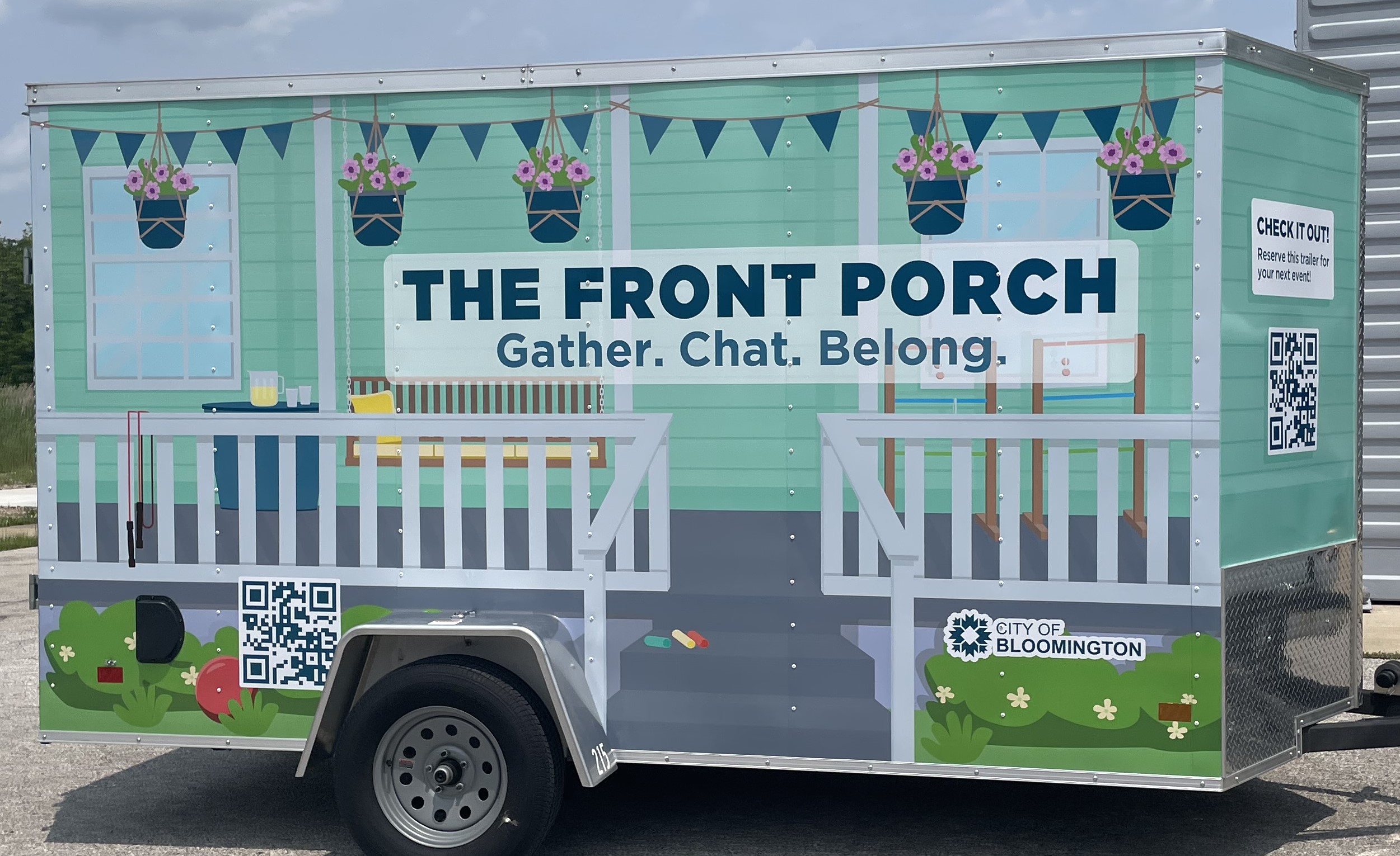 A cargo trailer decorated to look like a wraparound porch with a white railing, porch swing, and hanging flower baskets