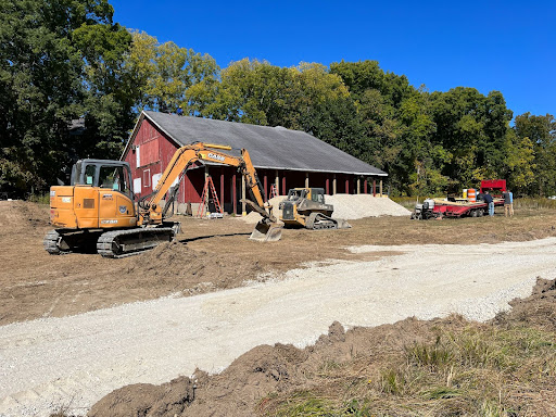 An excavator is parked on earth  between a graveled trail and a red barn