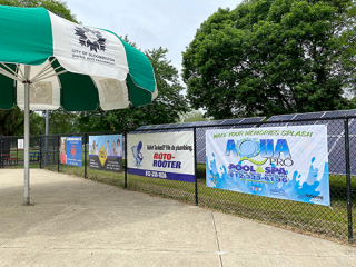 Colorful 8' x 4' banners from various businesses hanging on the chain link perimeter fence at Bryan Park Pool.