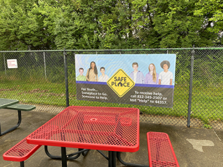 Photo of bright red outdoor picnic table at Mills Pool, with colorful 8' x 4' Youth Services Bureau sponsor banner hanging on the chain link perimeter fence.