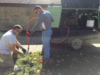 Two men, one kneeling and one standing, use an orange vacuum hose to clear water pipes.
