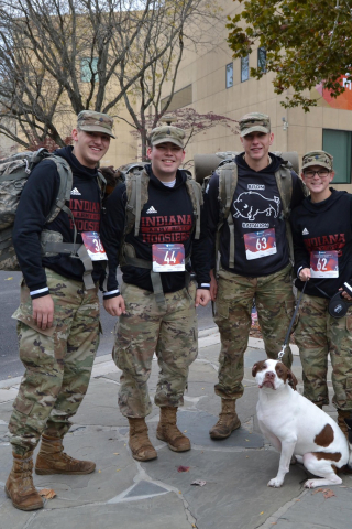 four service members wearing runner's bibs for Veterans 5K. One member holding a leash to white and brown dog.