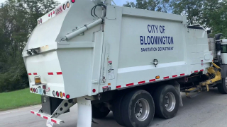 A white garbage truck passing by a residential street with "City of Bloomington Sanitation Department" written on its side in blue letters 