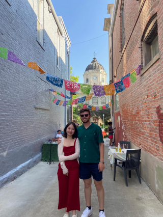 Two people smiling and facing the camera as they stand in the middle of an activated alley. The activated alley features strung papel picado, a live muralist, and seating. The Monroe County courthouse can be seen in the background.