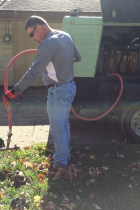 Two men, one kneeling and one standing, use an orange vacuum hose to clear water pipes.