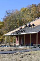 Photo shows a red barn at Goat Farm Park, with six people sitting on the roof and scraping off black shingles to reveal light colored decking beneath.