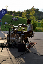Man sitting at drum set on Switchyard Park stage with green hills in the background