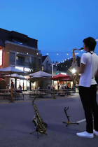 Young adult in white tshirt and dark pants plays saxophone and sunset on Kirkwood in front of residents/visitors using outdoor seating
